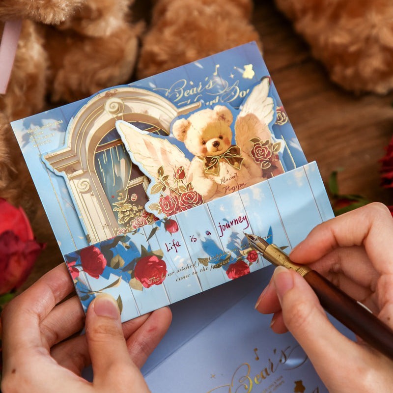 StationeryHaul angelic teddy bear pop-up card with wings and roses, blue sky background, gold accents.